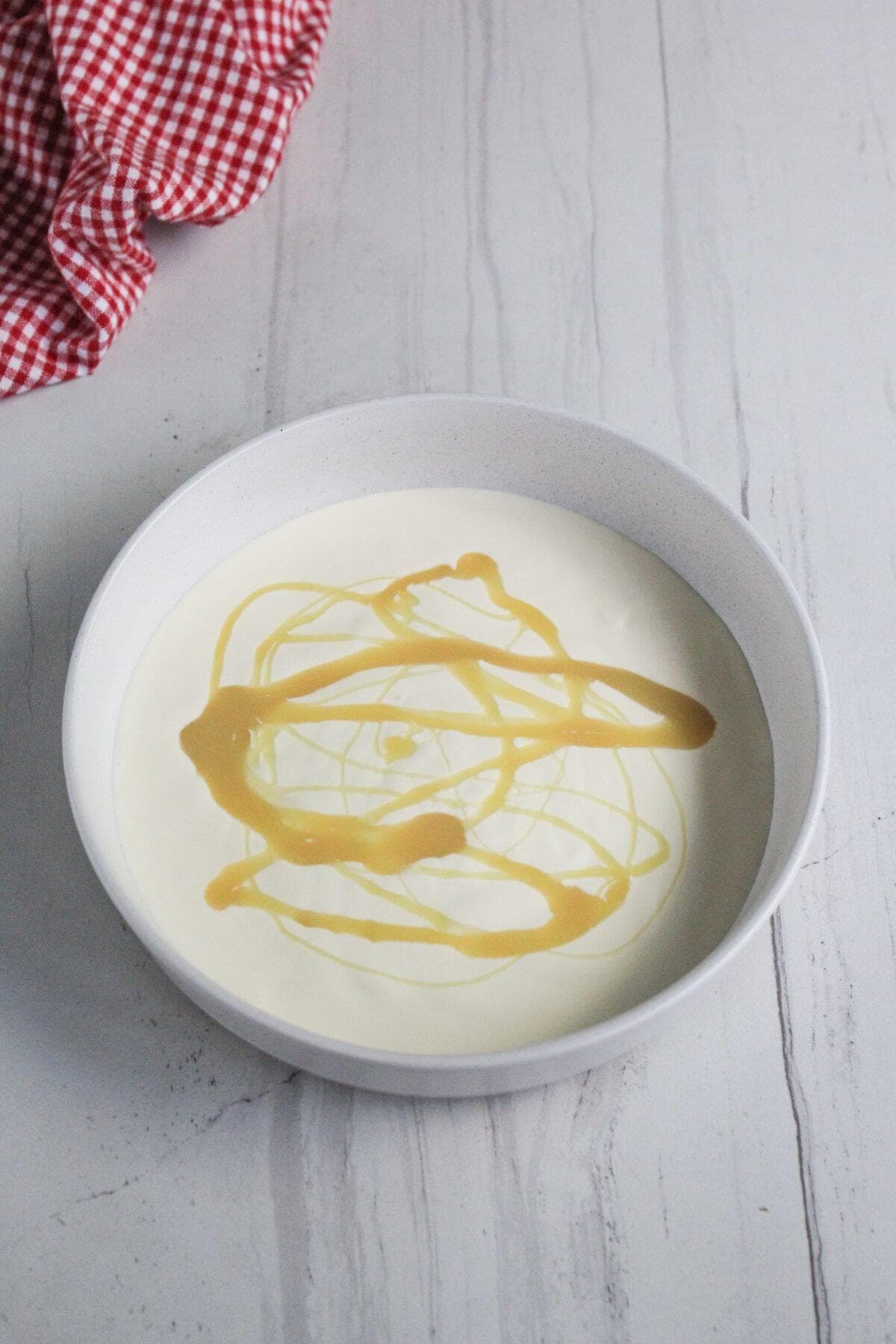 A white bowl filled with yogurt topped with a drizzle of honey sits on a white marble surface; a red and white checkered cloth is nearby.