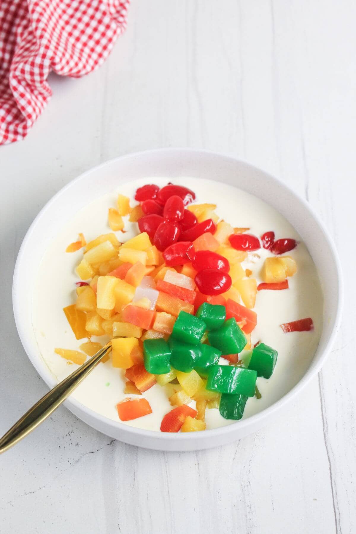 A white bowl filled with colorful fruit cocktail pieces and nata de coco in a white creamy liquid, with a gold spoon on the side.