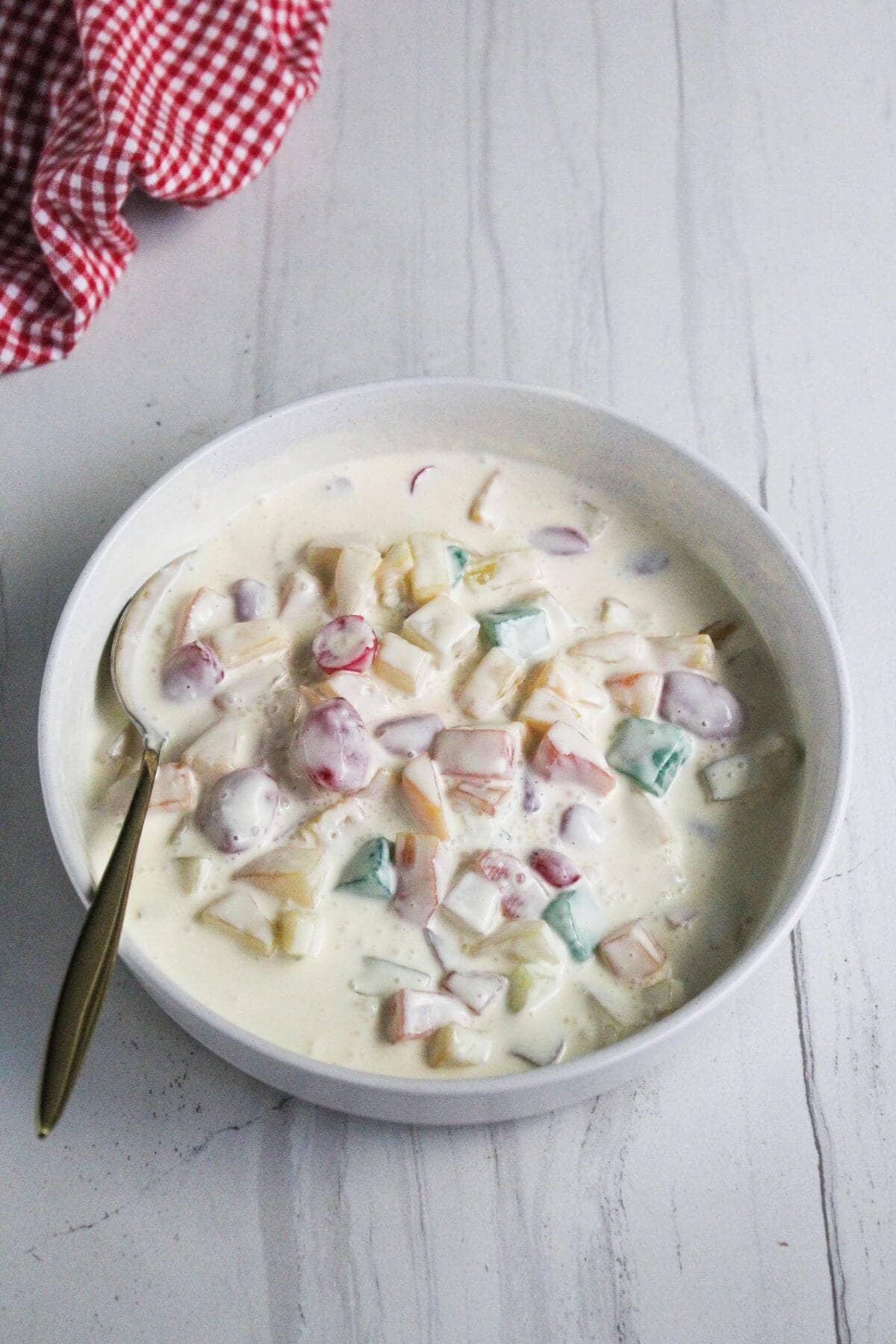 A bowl of fruit salad with colorful fruit pieces in creamy dressing, placed on a white surface with a spoon and a red cloth nearby.