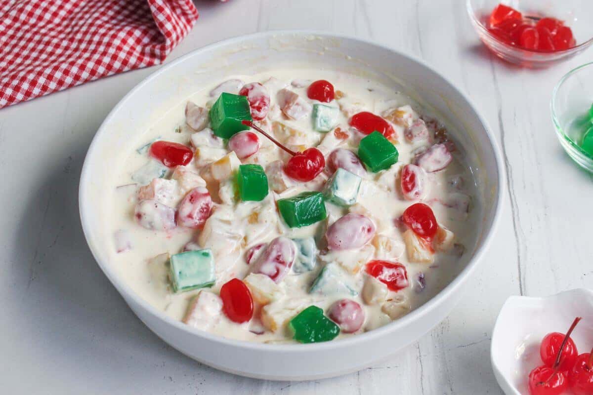 A bowl of fruit salad with colorful jelly cubes, cherries, and cream, on a white table with a red checkered cloth nearby.