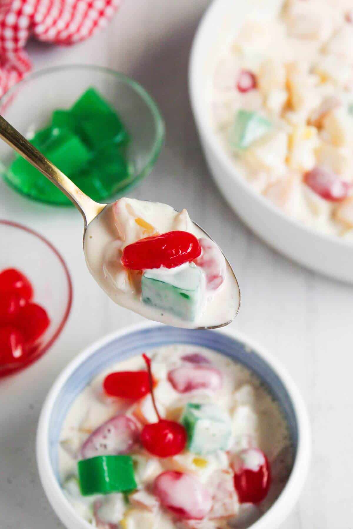 A spoonful of creamy fruit salad with red and green jelly cubes, cherries, and mixed fruit over a bowl, with extra jelly and cherries in small bowls nearby.