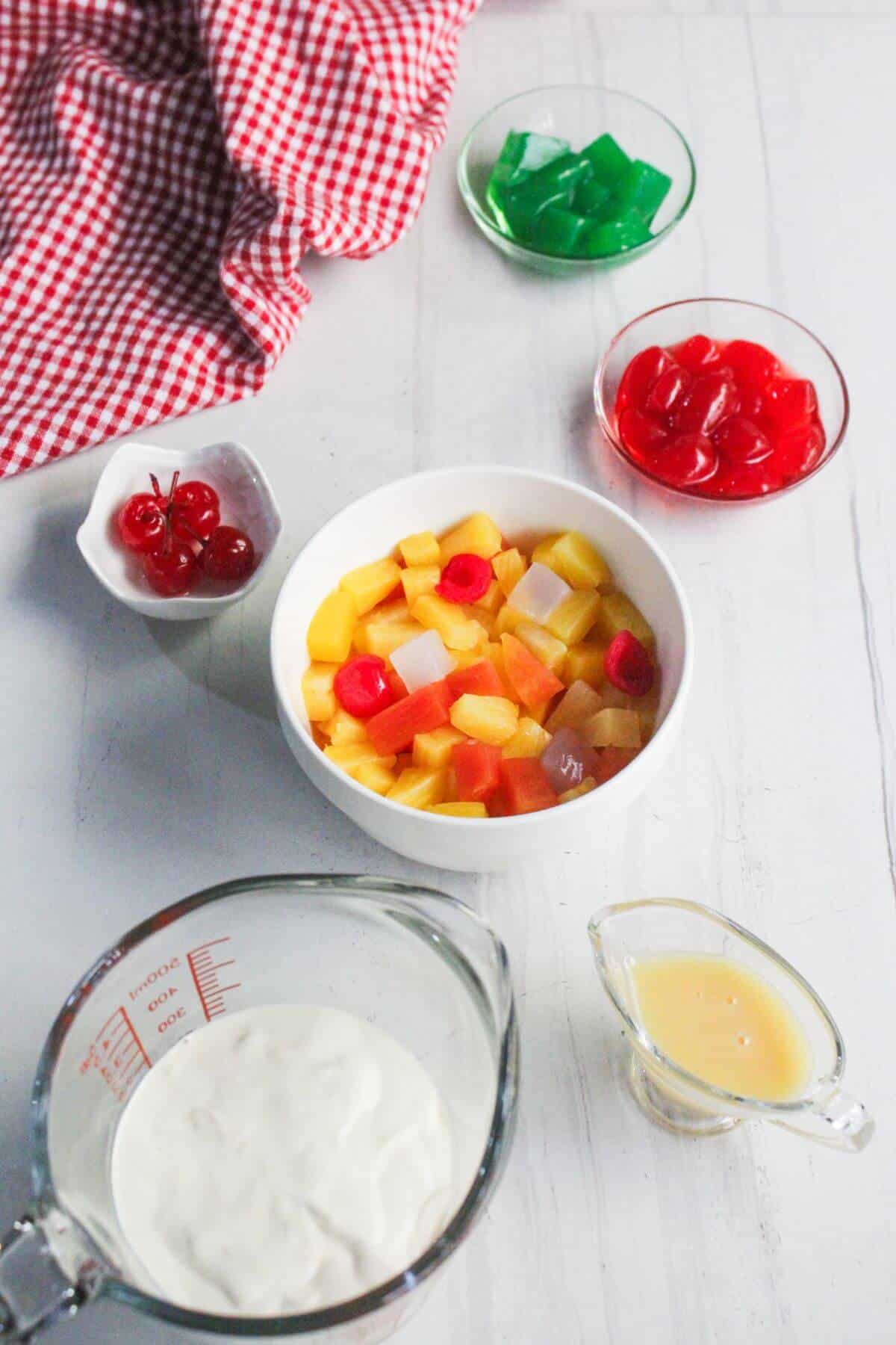 Assorted fruits, cherries, green gelatin, red gelatin, creamy mixture, and liquid in bowls and a measuring cup on a white surface with a red-checkered cloth.