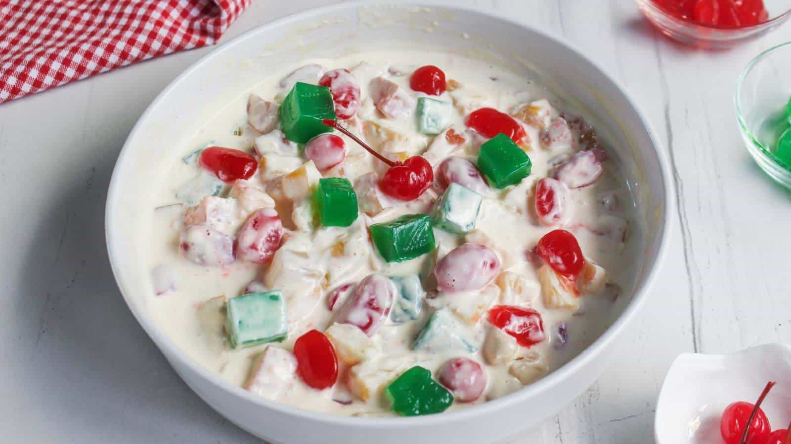 A white bowl filled with fruit salad in creamy sauce, topped with green gelatin cubes and maraschino cherries, sits on a white surface.