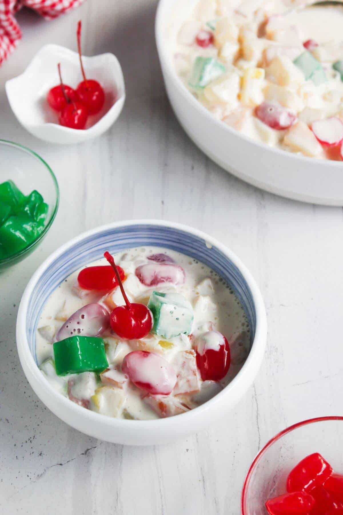 A bowl of fruit salad with colorful gelatin cubes, grapes, and cherries in a creamy sauce, with more fruit salad and cherries in the background.