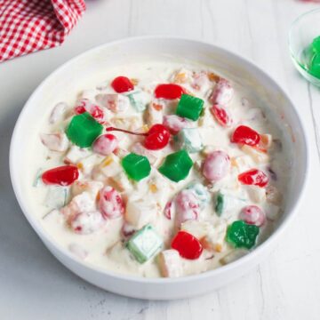 A white bowl filled with fruit salad in creamy sauce, topped with red cherries, green gelatin cubes, and assorted fruit pieces on a white surface.