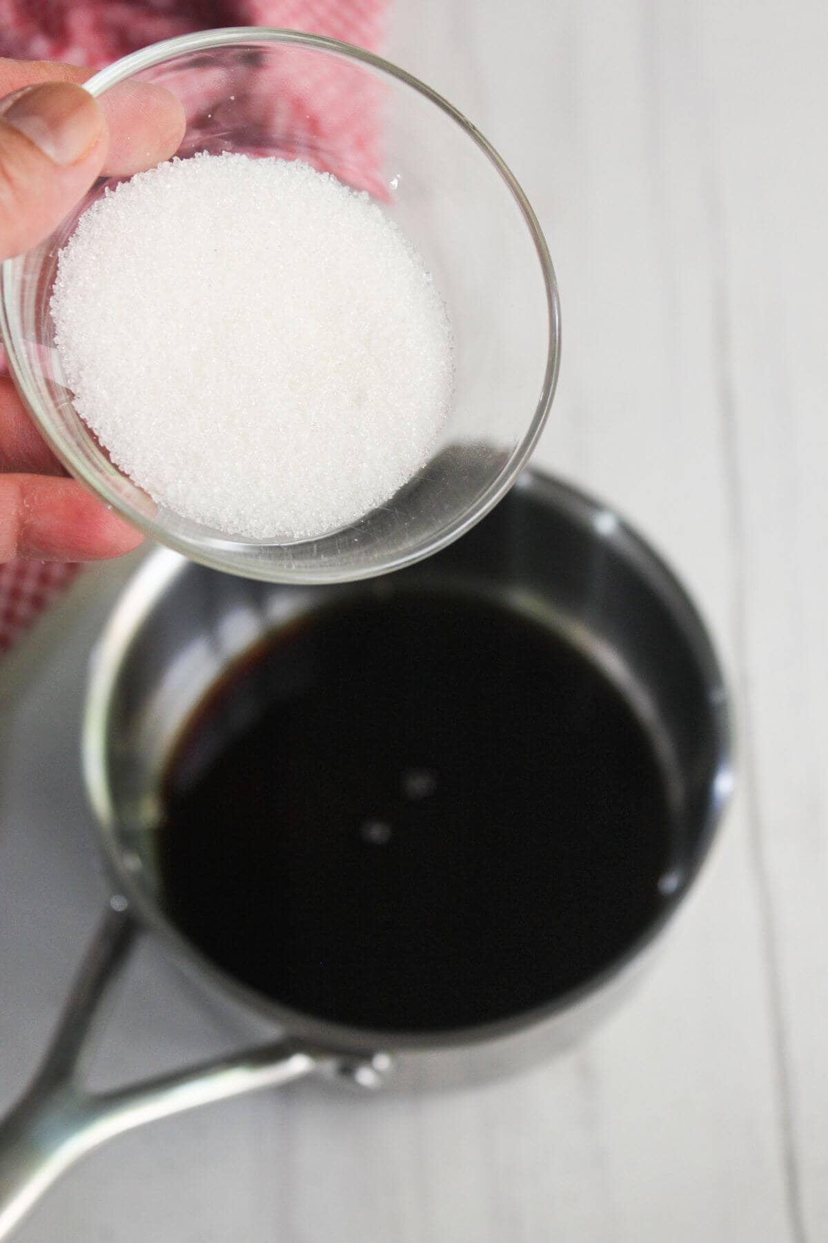 A hand holds a small glass bowl of white granulated sugar above a saucepan containing a dark liquid on a white surface.