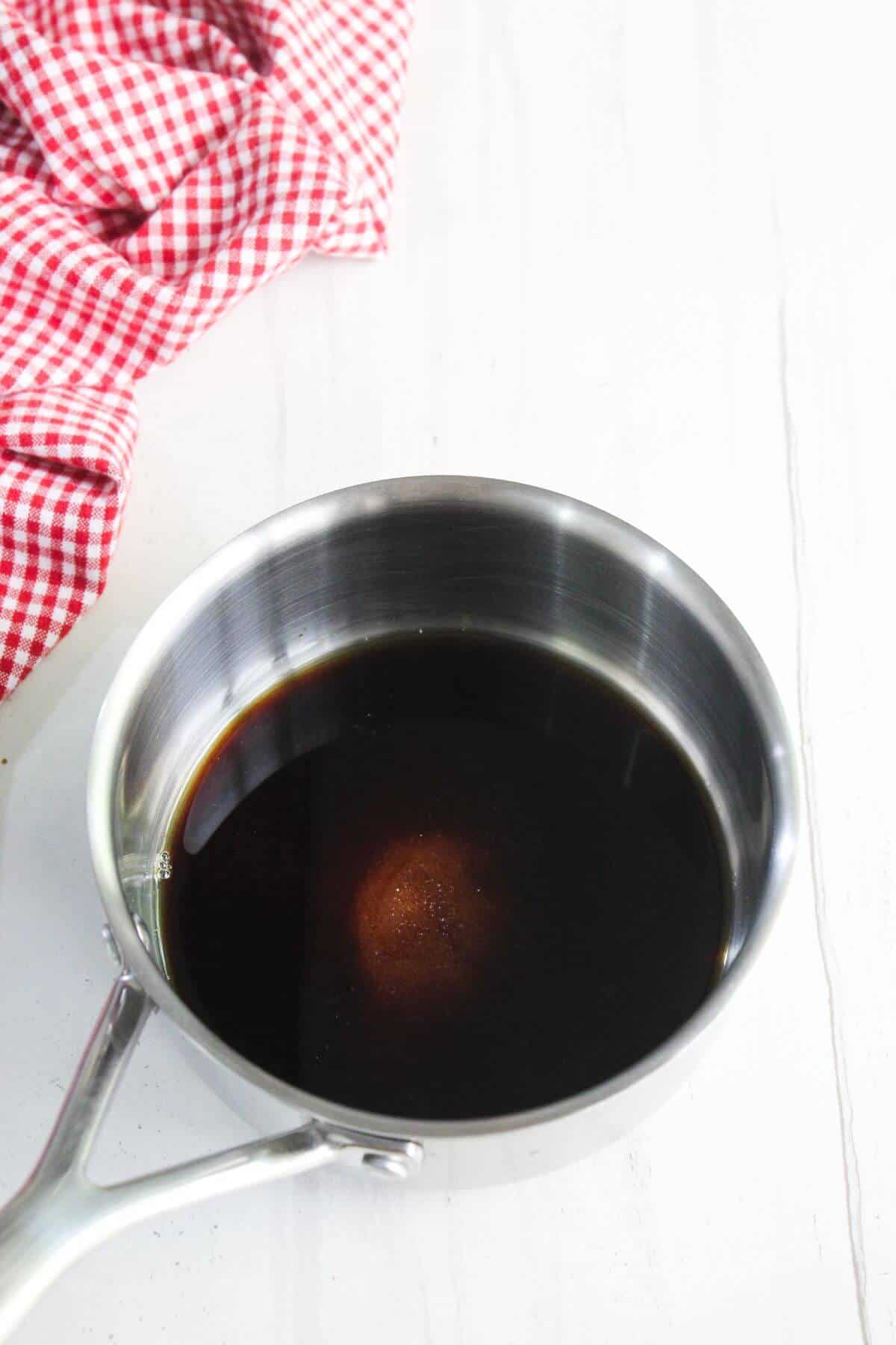 A stainless steel saucepan containing a dark liquid sits on a white surface next to a red and white checkered cloth.