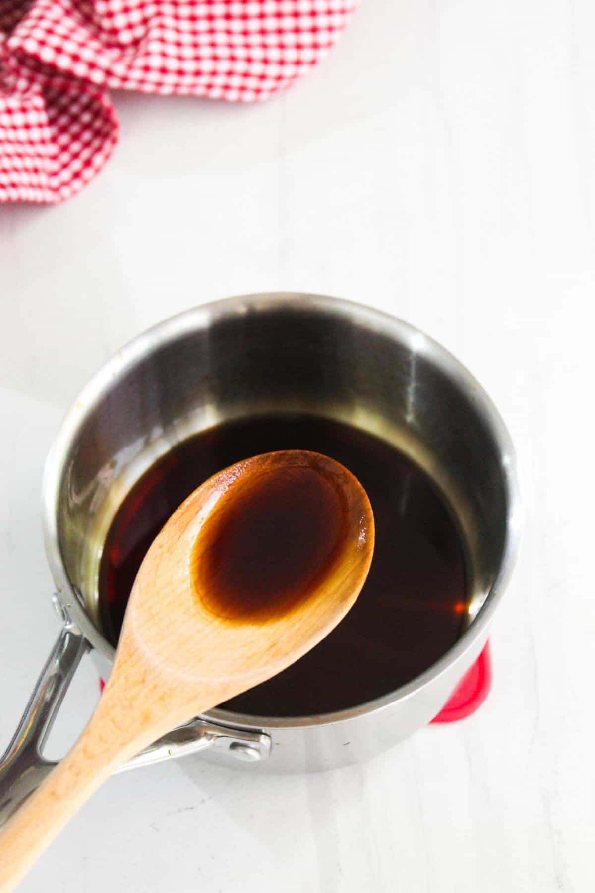 A wooden spoon with dark liquid over a saucepan containing the same liquid, placed on a white surface with a red checkered cloth in the background.