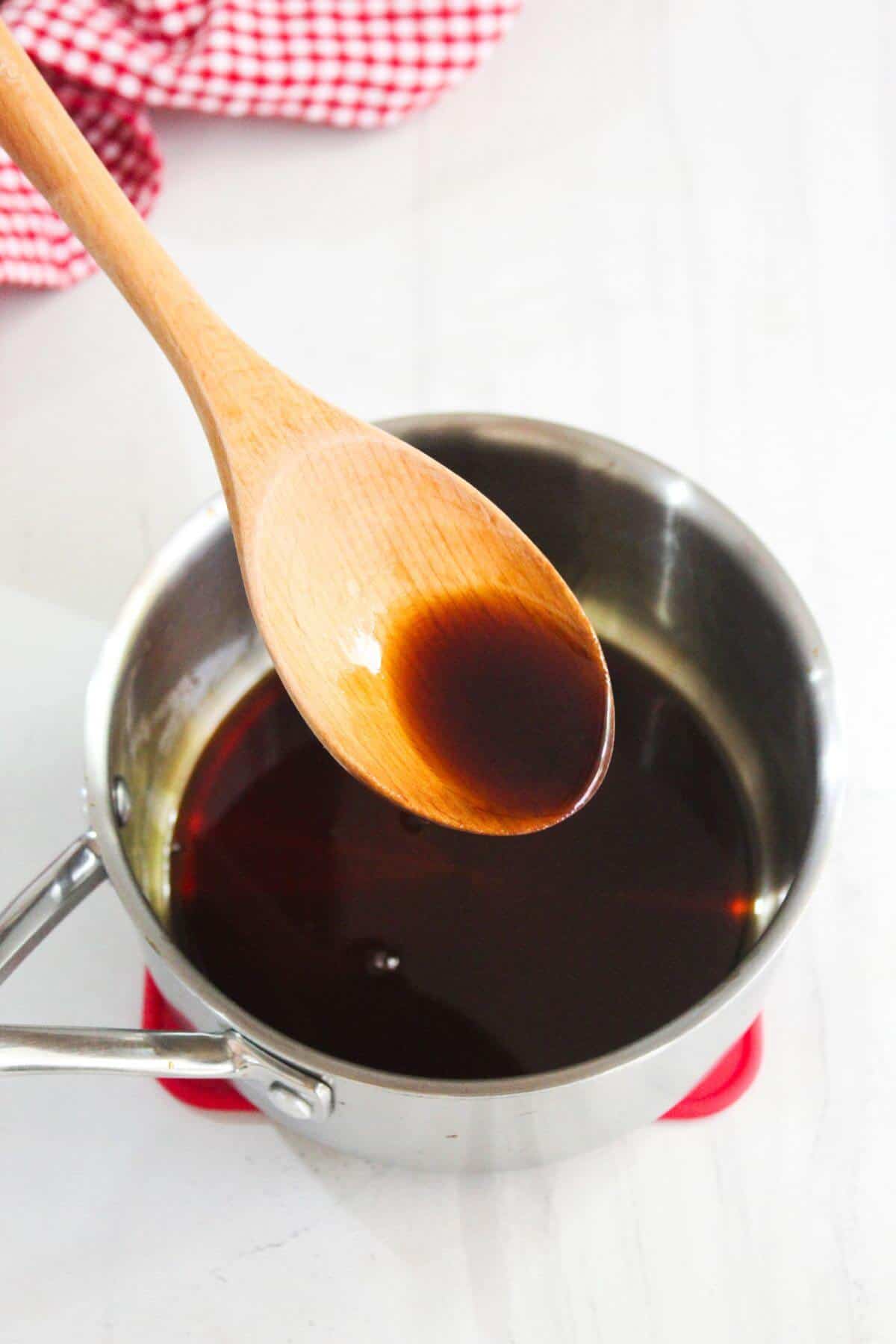 A wooden spoon holds a small amount of dark brown liquid above a stainless steel saucepan filled with the same liquid on a white surface.