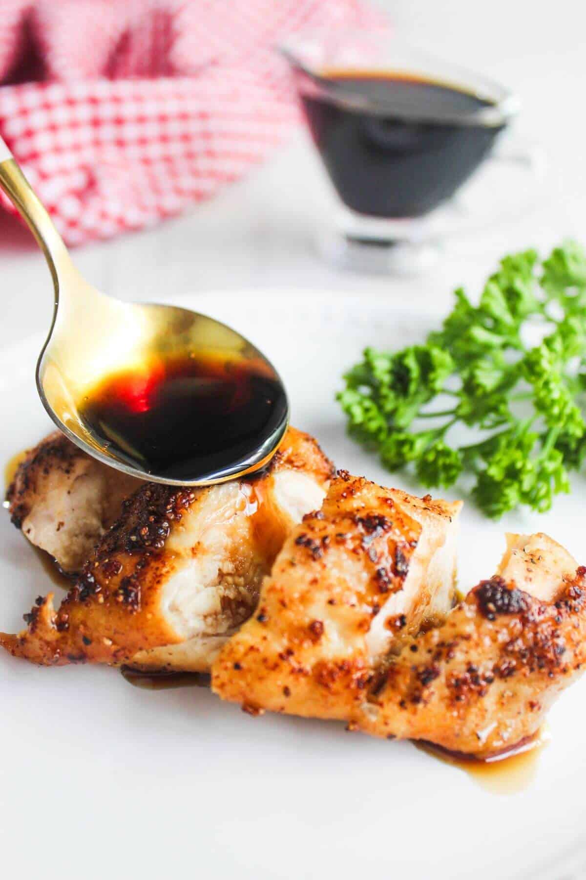 A golden spoon drizzles a dark sauce over sliced, cooked chicken breast on a white plate, garnished with fresh parsley; a sauce container and red cloth are in the background.