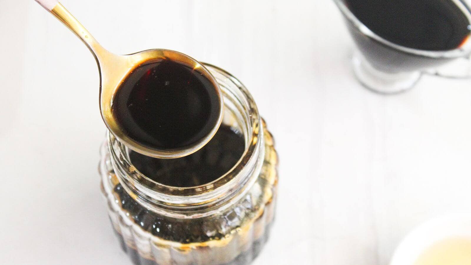 A spoon holds dark teriyaki sauce over an open glass jar filled with more teriyaki sauce; a small sauce pitcher is visible in the background.