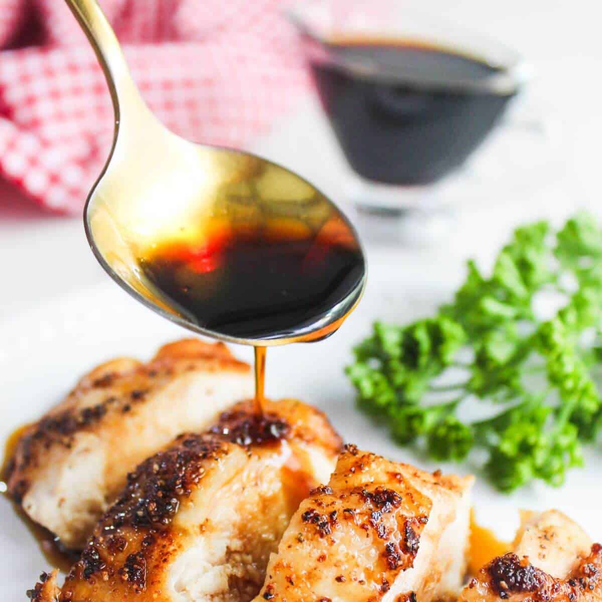 Sliced grilled chicken is topped with a dark sauce from a gold spoon, with a sprig of parsley and a sauce dish in the background.