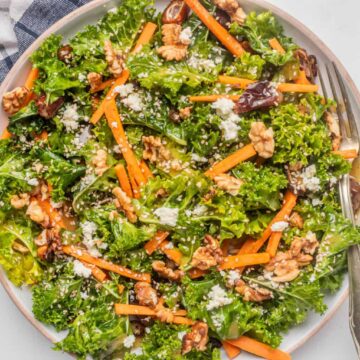 A plate of kale salad topped with carrot sticks, walnuts, crumbled cheese, and sesame seeds, with a fork on the side.