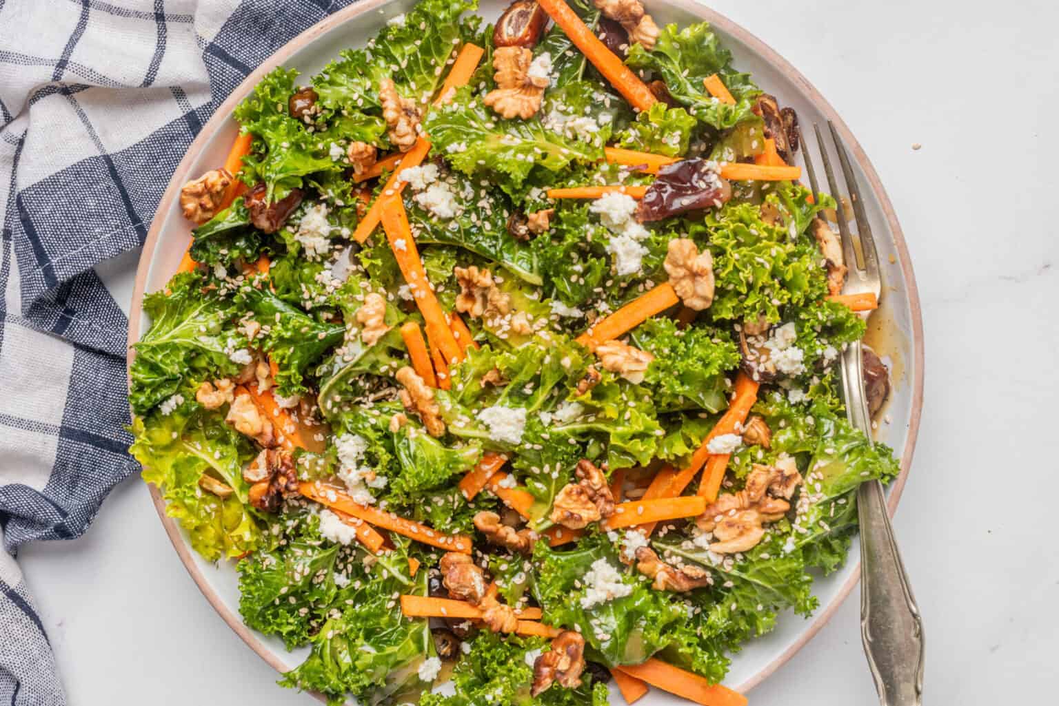 A plate of kale salad topped with carrot sticks, walnuts, crumbled cheese, and sesame seeds, with a fork on the side.