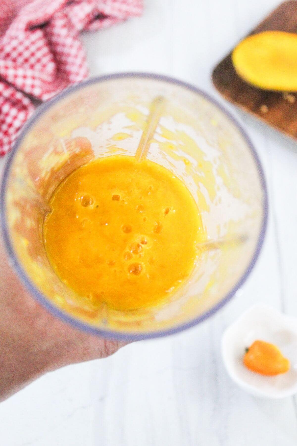 A hand holds a blender cup filled with a thick, yellow-orange mixture. A red cloth, sliced mango, and a small pepper are visible in the background.