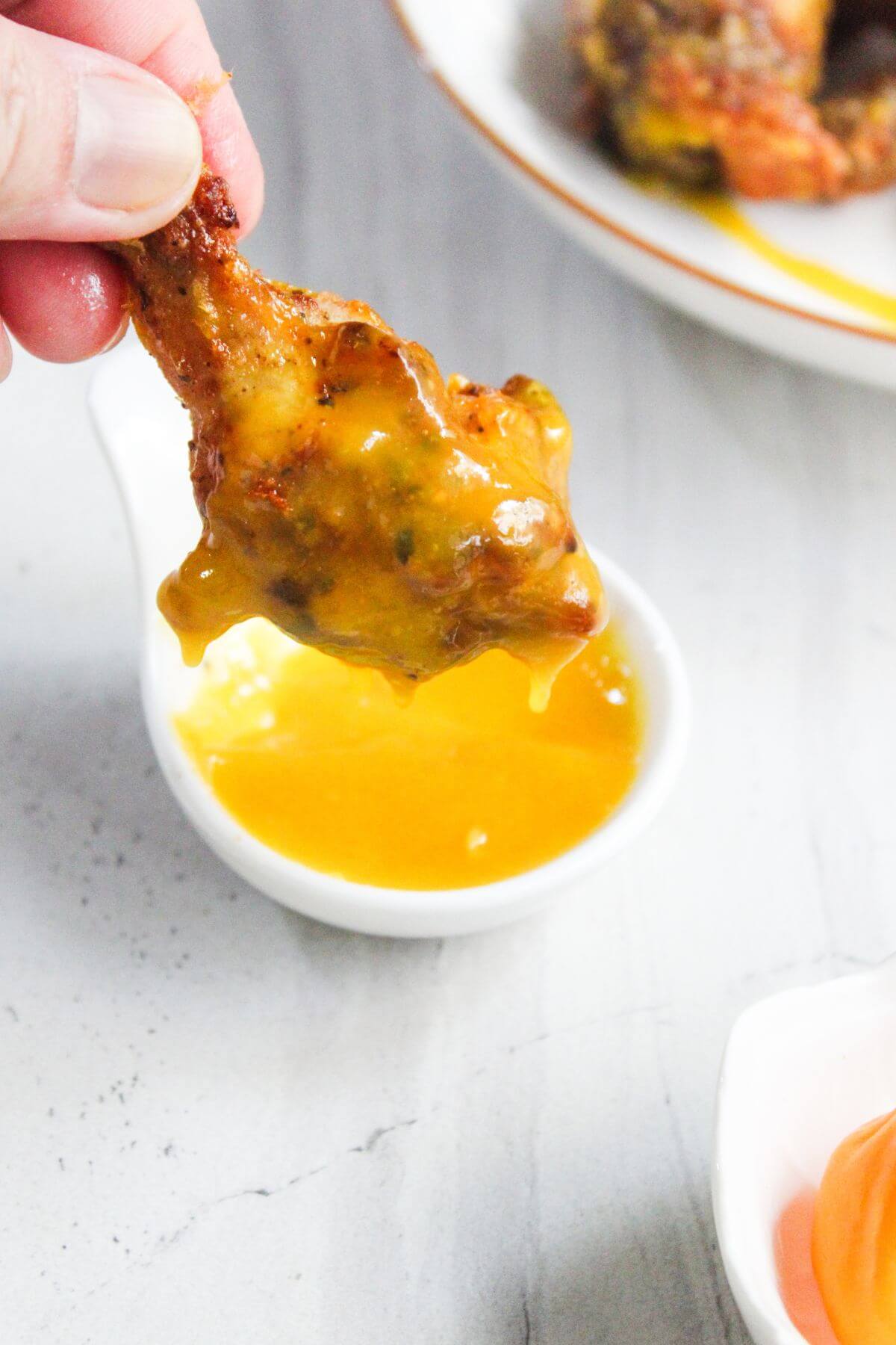 A hand dips a chicken wing into a small dish of yellow sauce, with sauce dripping from the wing.