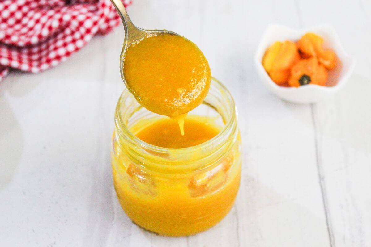 A spoon holds yellow hot sauce over an open glass jar, with a small bowl of orange peppers and a red checkered cloth in the background.