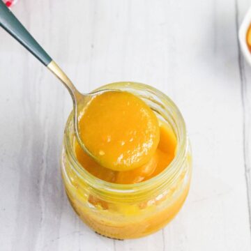A spoon scooping thick, yellow mango puree from a glass jar on a light-colored surface.