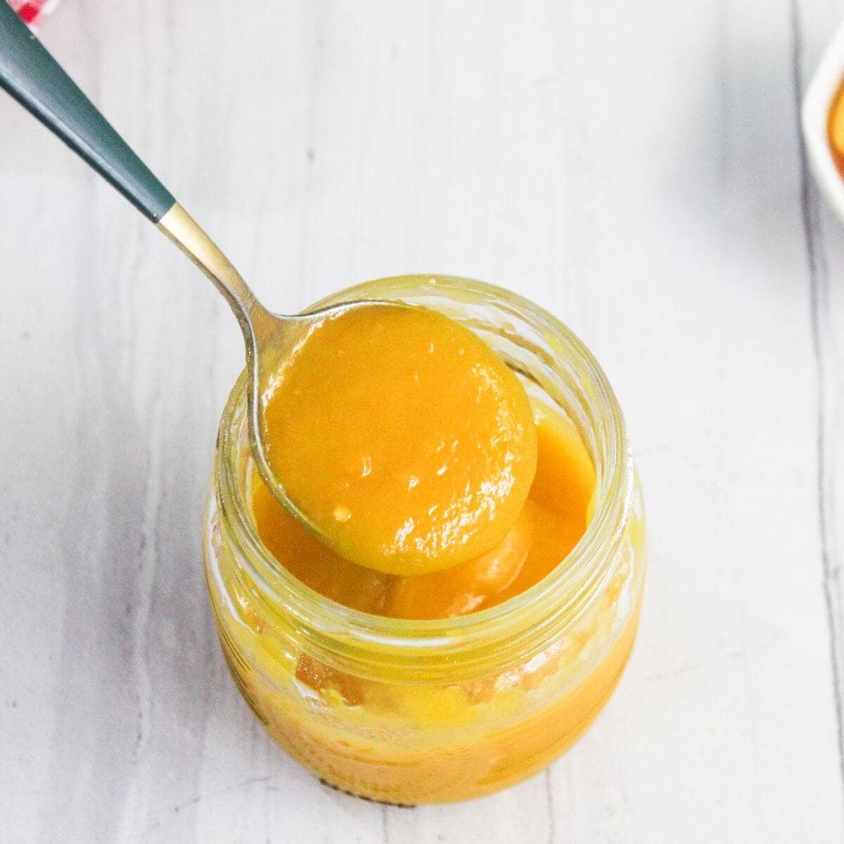 A spoon scooping thick, yellow mango puree from a glass jar on a light-colored surface.