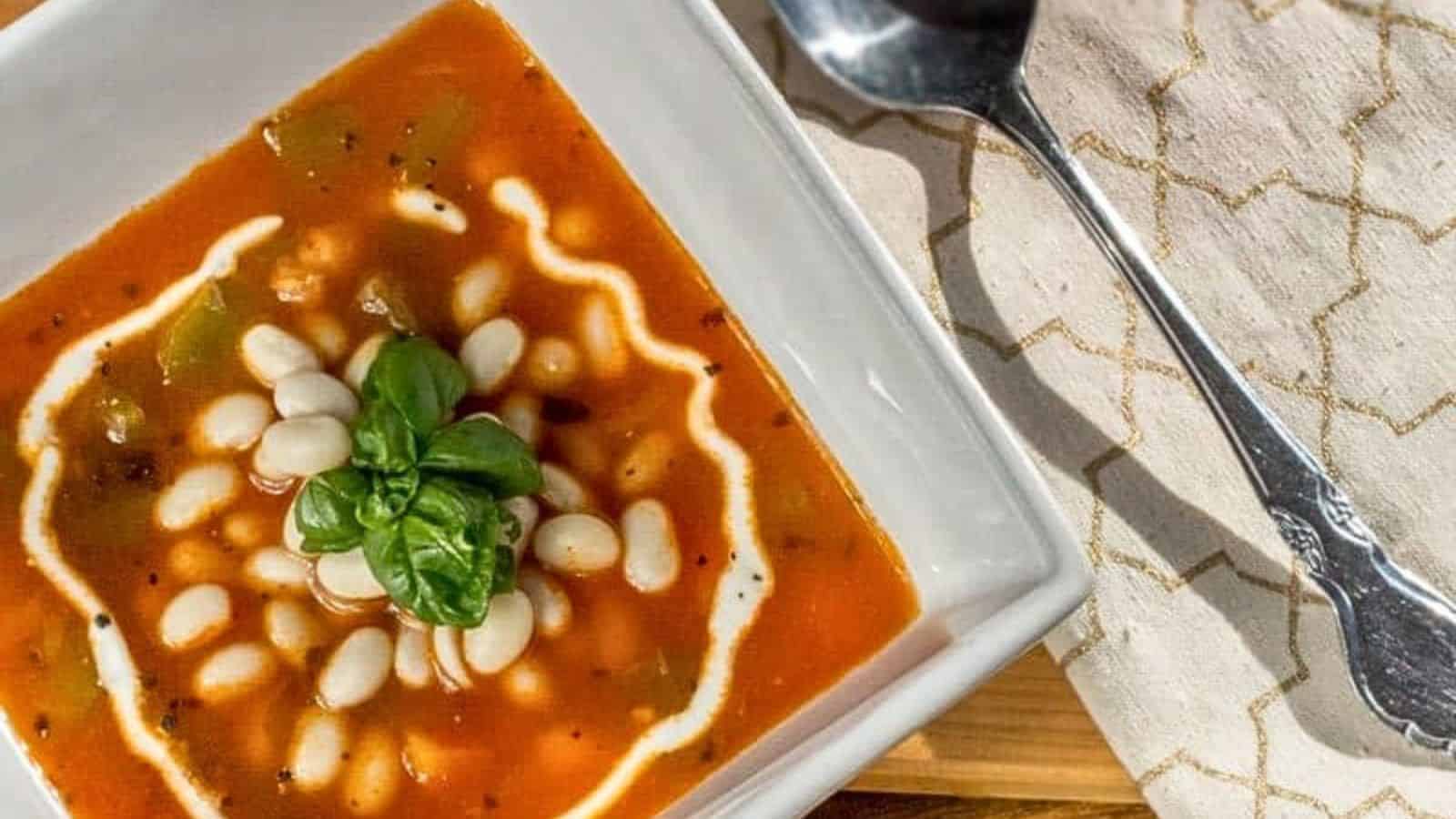 A bowl of tomato-based soup garnished with white beans, cream, and fresh basil, placed on a napkin beside a silver spoon.