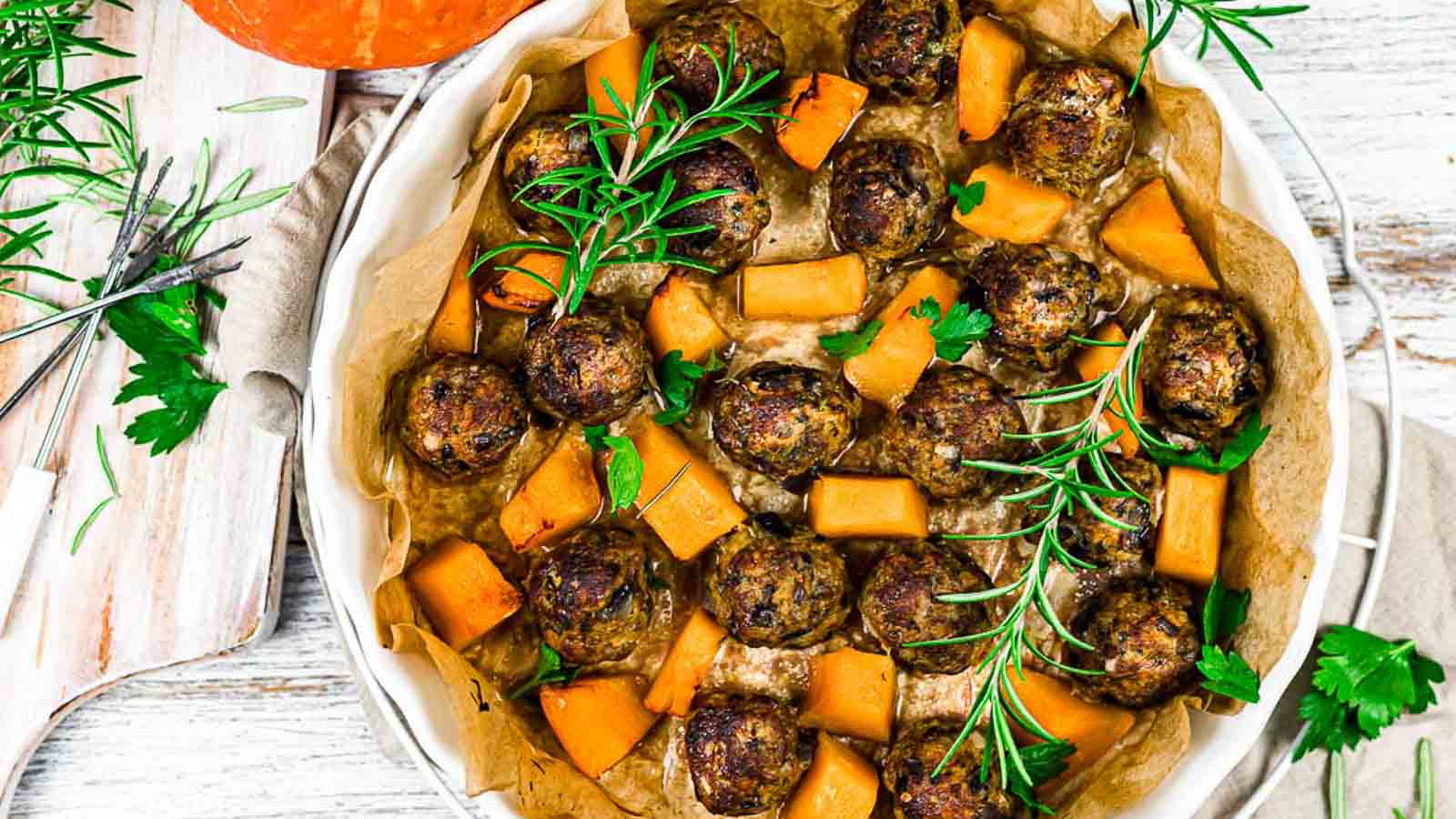 A round baking dish lined with parchment paper, filled with meatballs, cubed squash, fresh rosemary, and parsley.