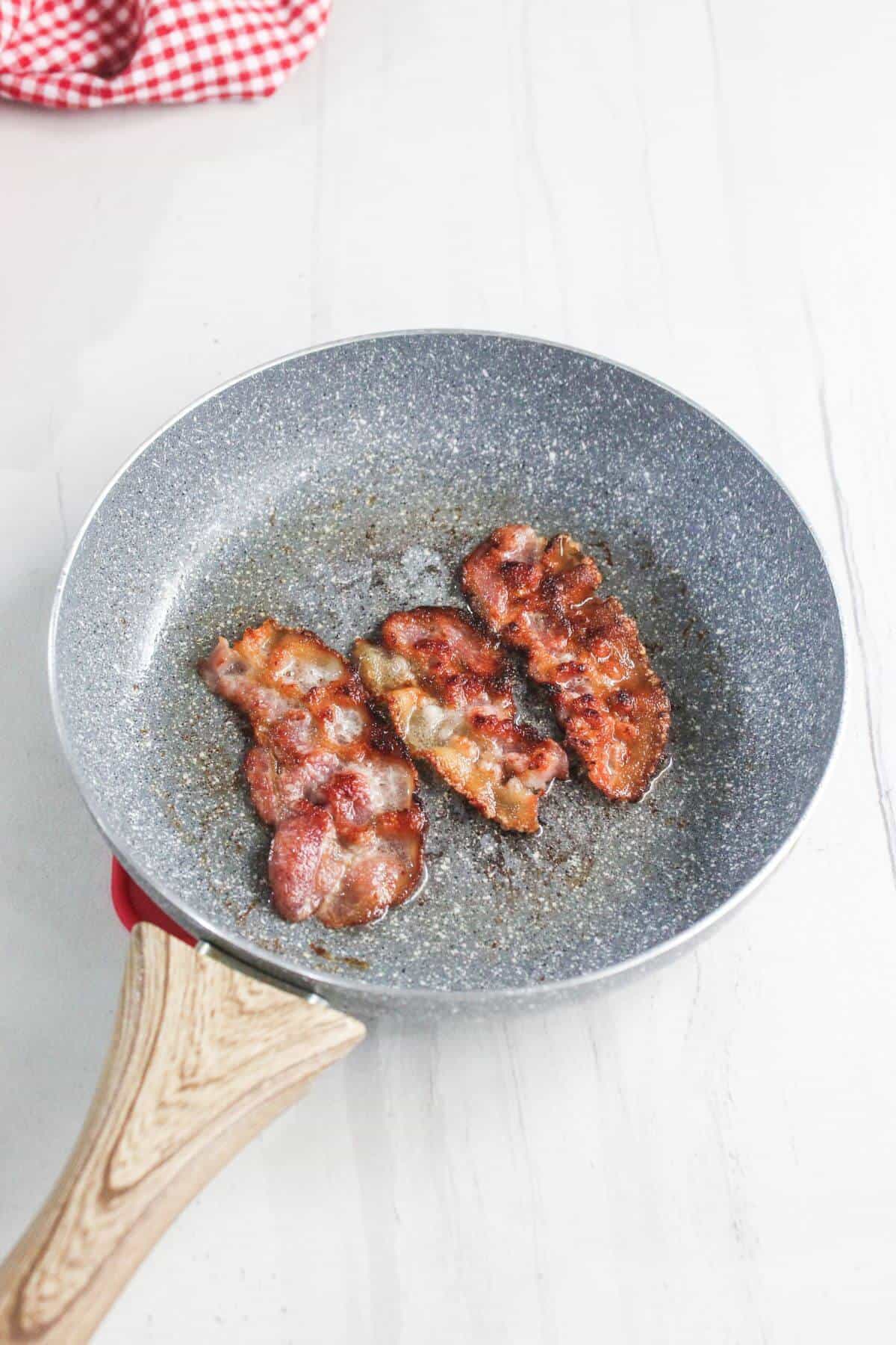 Three strips of cooked bacon sit in a frying pan with some grease on a light-colored surface.