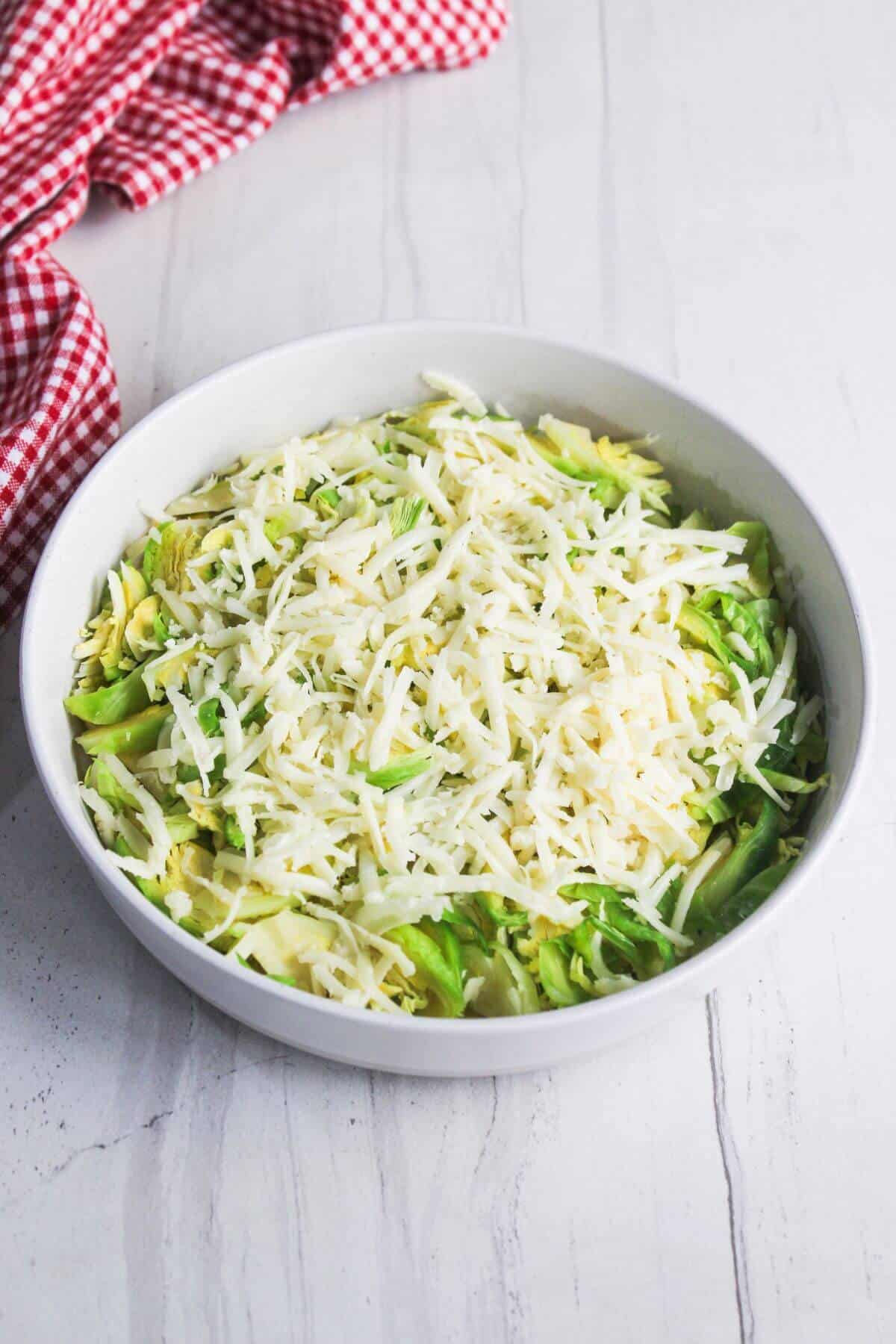 A white bowl filled with shredded cheese on top of sliced Brussels sprouts sits on a light marble surface next to a red and white checkered cloth.