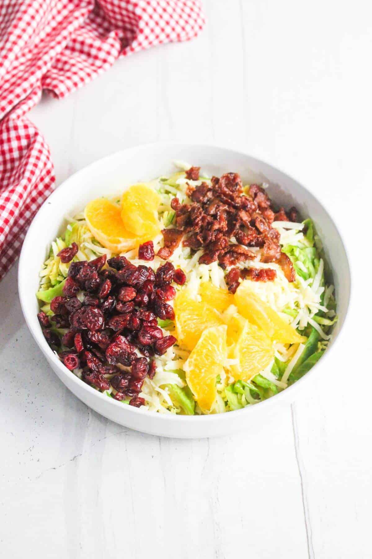 A white bowl filled with shredded cabbage, orange slices, dried cranberries, and chopped bacon, placed on a white surface beside a red and white checkered cloth.