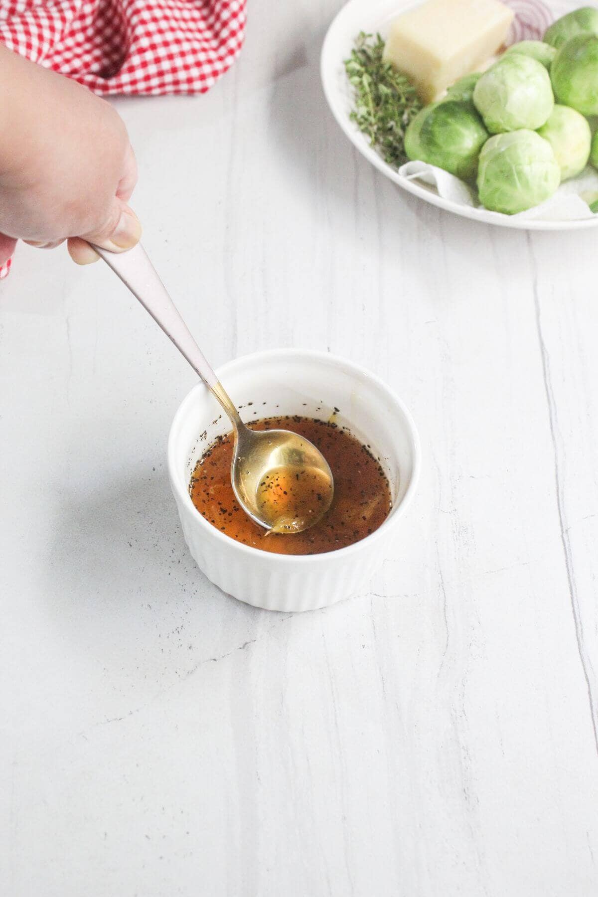A hand holds a spoon in a ramekin containing a seasoned liquid, with a plate of Brussels sprouts, cheese, and herbs in the background.