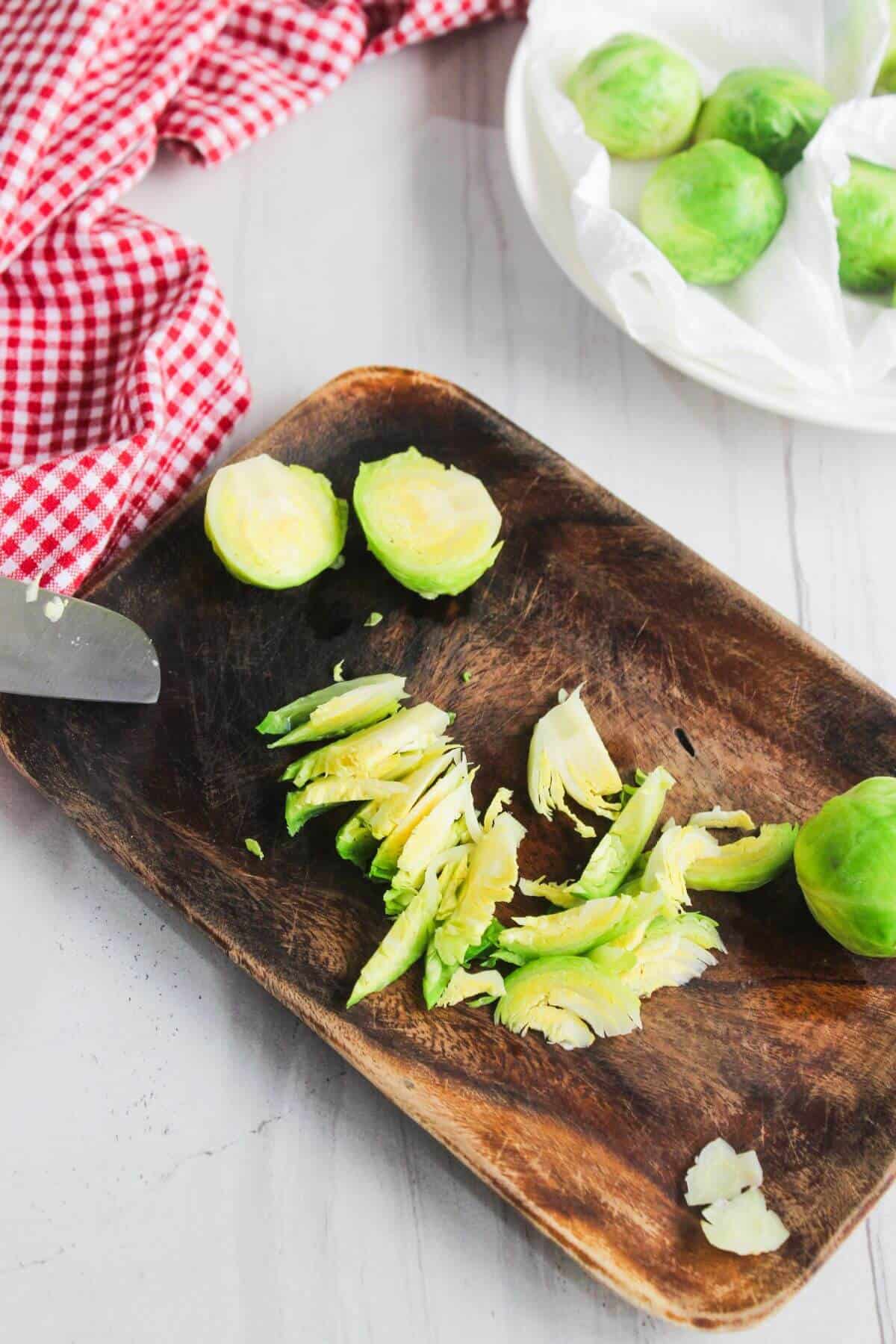 A wooden board with halved and sliced Brussels sprouts, a knife, and a plate with whole Brussels sprouts on a white surface with a red checkered cloth.