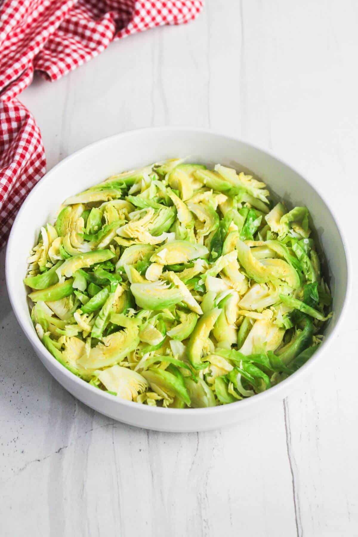 A white bowl filled with sliced raw Brussels sprouts sits on a light marble surface near a red and white checkered cloth.