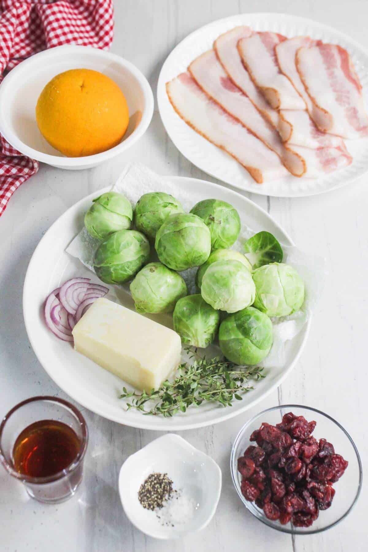 A plate of fresh Brussels sprouts, butter, sliced red onion, thyme, sliced bacon, an orange, dried cranberries, black pepper, and a small glass of maple syrup on a white surface.