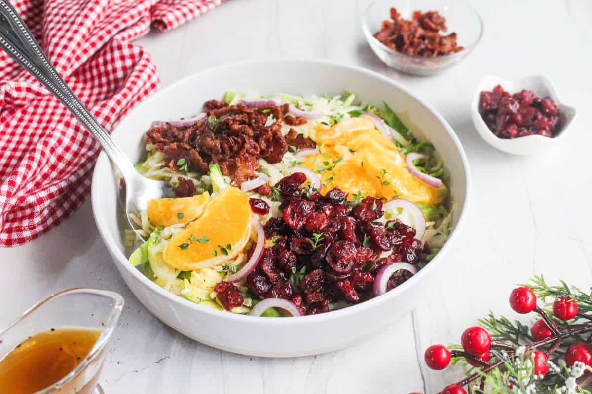 A bowl of salad with orange slices, dried cranberries, red onions, and chopped bacon, with a fork and small bowls of cranberries and bacon on the side.