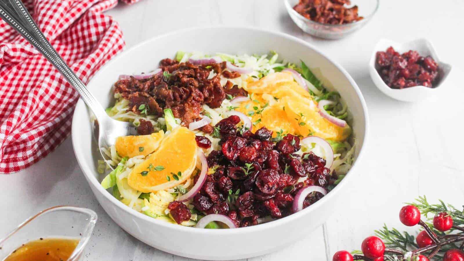A white bowl filled with salad containing orange slices, dried cranberries, red onion, shredded greens, and bacon bits, with a fork resting inside. Small bowls of toppings and dressing nearby.