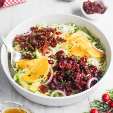 A salad in a white bowl featuring orange slices, dried cranberries, red onion rings, and bacon bits on a bed of lettuce. A fork is placed in the bowl.