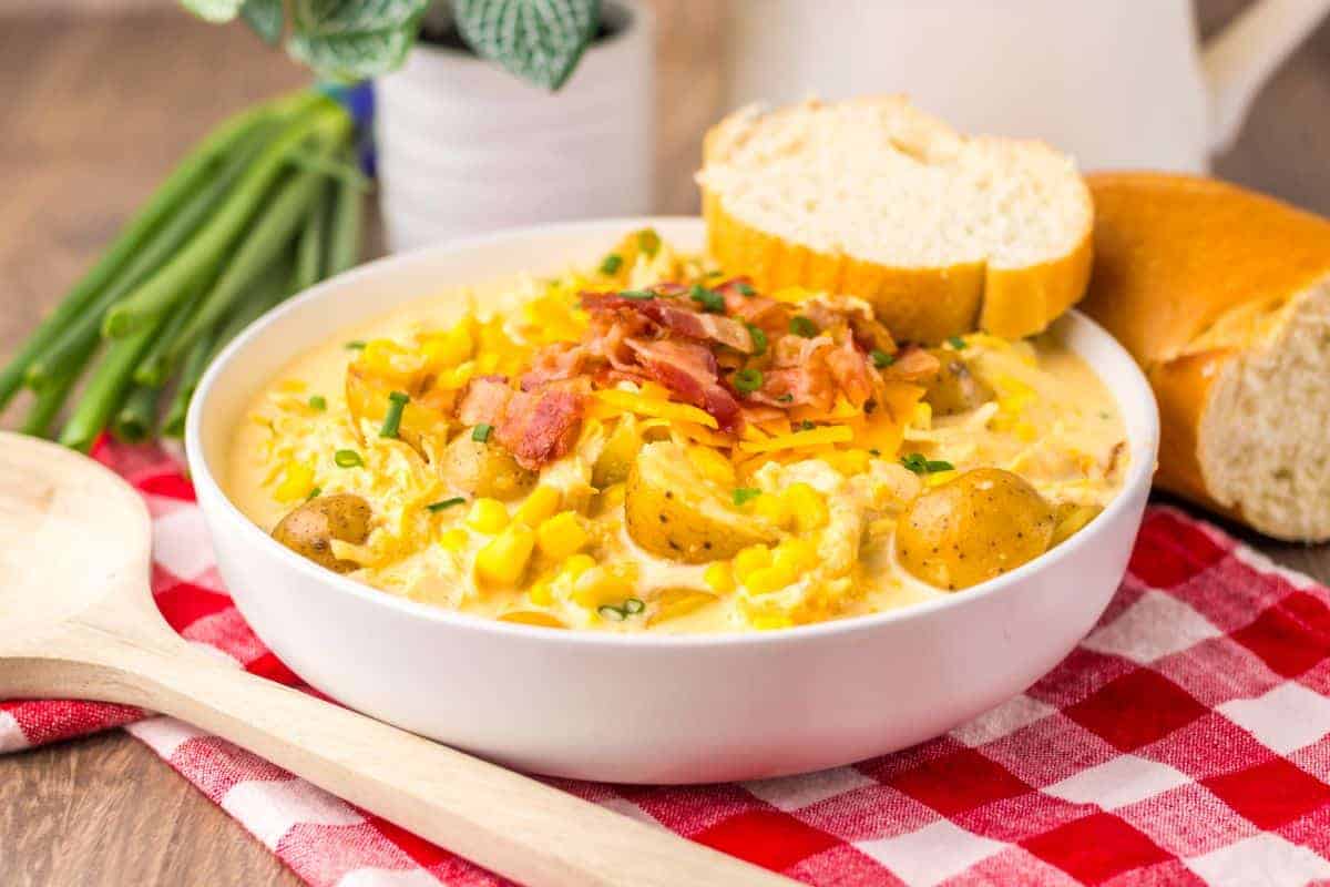 A bowl of potato and corn chowder topped with shredded cheese and bacon, served with slices of bread on a red checkered cloth.