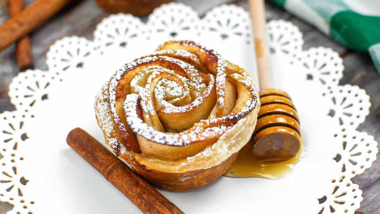 A pastry shaped like a rose dusted with powdered sugar on a white plate, with a honey dipper, honey, and a cinnamon stick beside it.