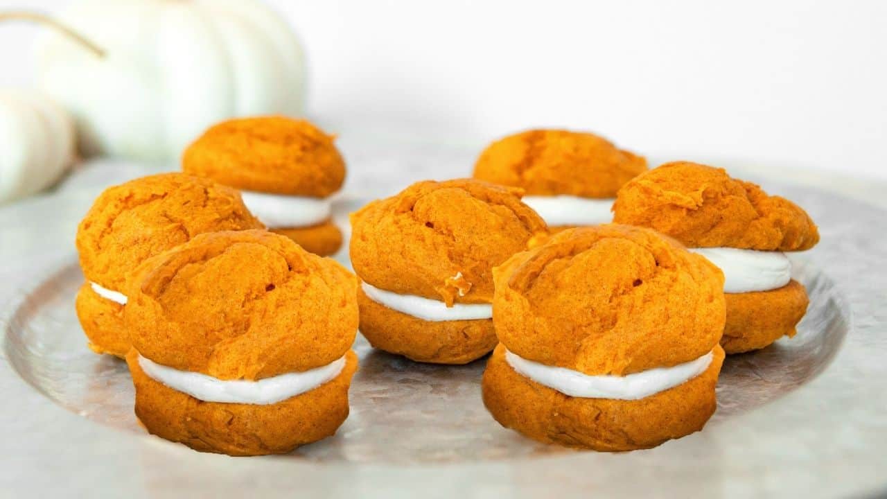Six pumpkin whoopie pies with white cream filling are arranged on a metallic tray. White pumpkins are blurred in the background.