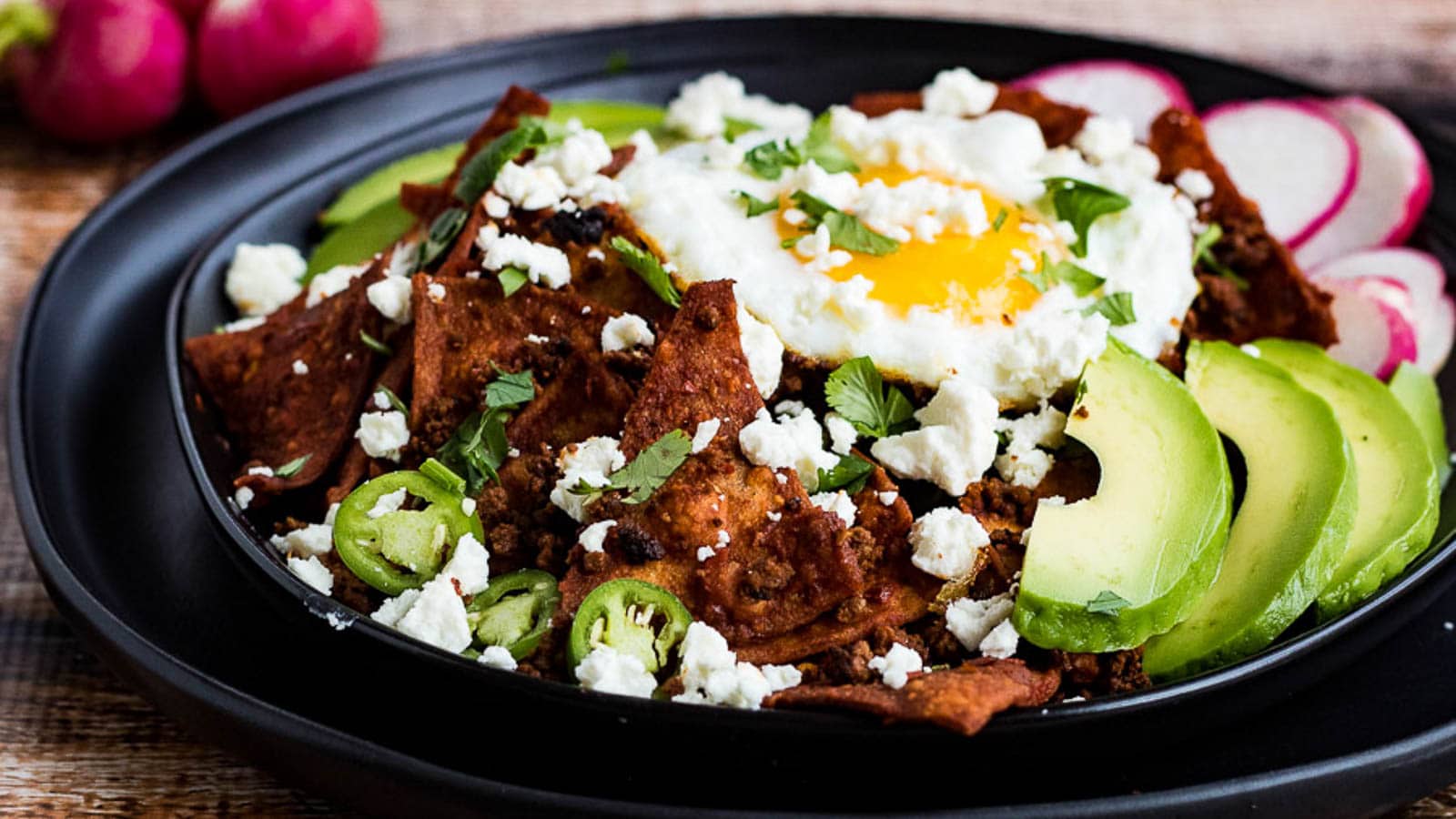 A plate of chilaquiles topped with a fried egg, avocado slices, sliced radishes, jalapeños, cilantro, and crumbled cheese on a black plate.