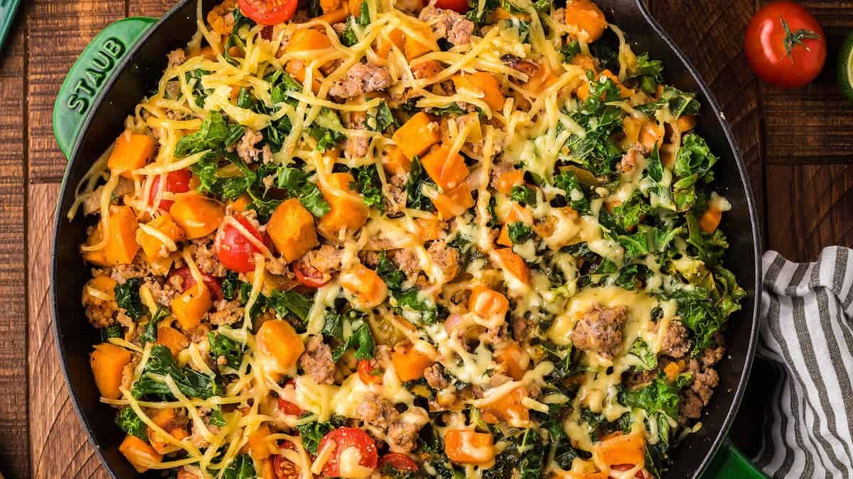 A skillet filled with a baked mixture of ground meat, leafy greens, chopped tomatoes, cubed sweet potatoes, and melted cheese.
