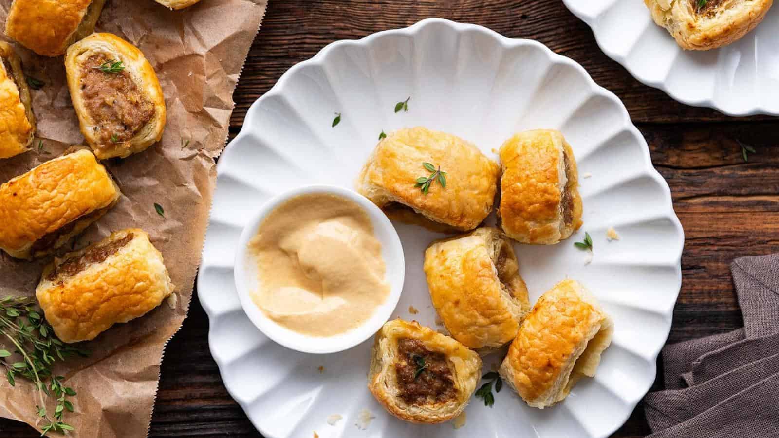 A white plate with several sausage rolls—classic savory puff pastry recipes—alongside a small bowl of dipping sauce, with more sausage rolls on brown parchment paper beside the plate.