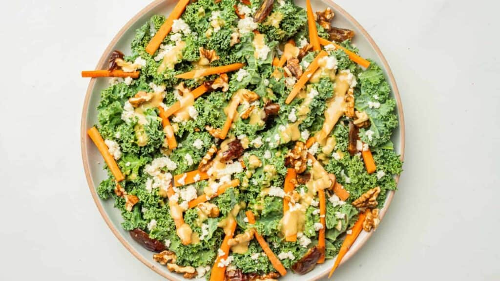 Plate of kale salad topped with carrot sticks, walnuts, crumbled cheese, dates, and a light drizzle of dressing on a white background.