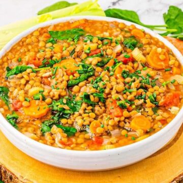 A bowl of lentil soup with chopped carrots, spinach, and herbs, surrounded by fresh vegetables like leeks, carrots, garlic, and spinach on a wooden board.