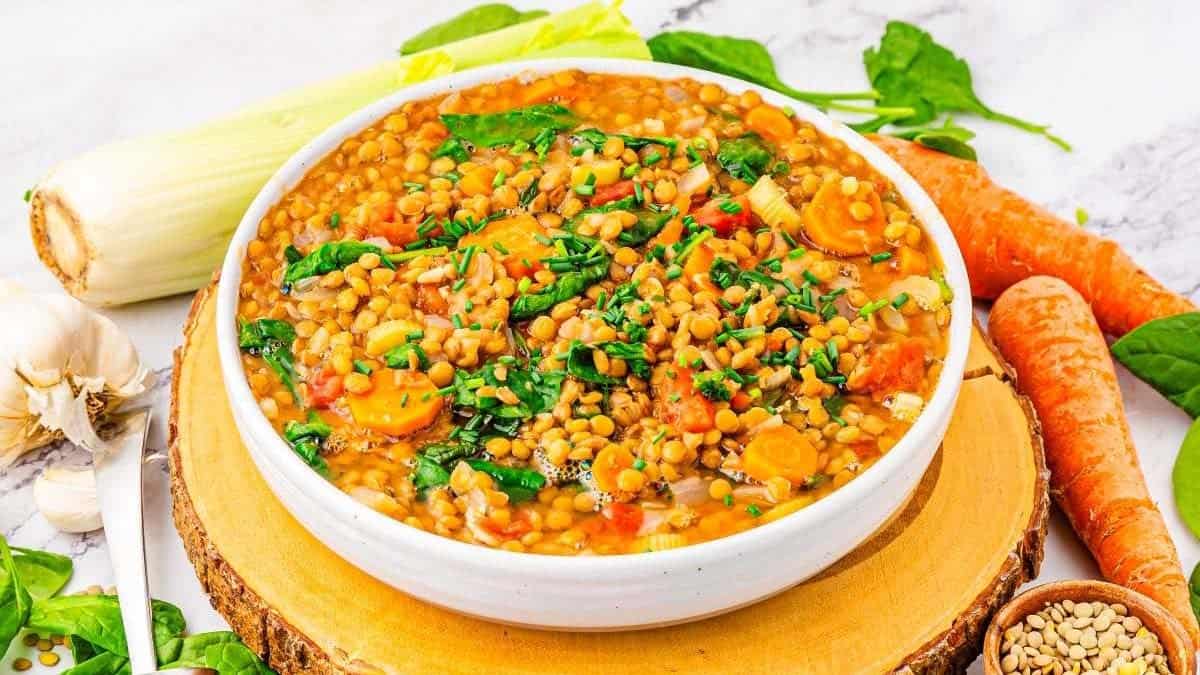 A bowl of lentil soup with chopped carrots, spinach, and herbs, surrounded by fresh vegetables like leeks, carrots, garlic, and spinach on a wooden board.