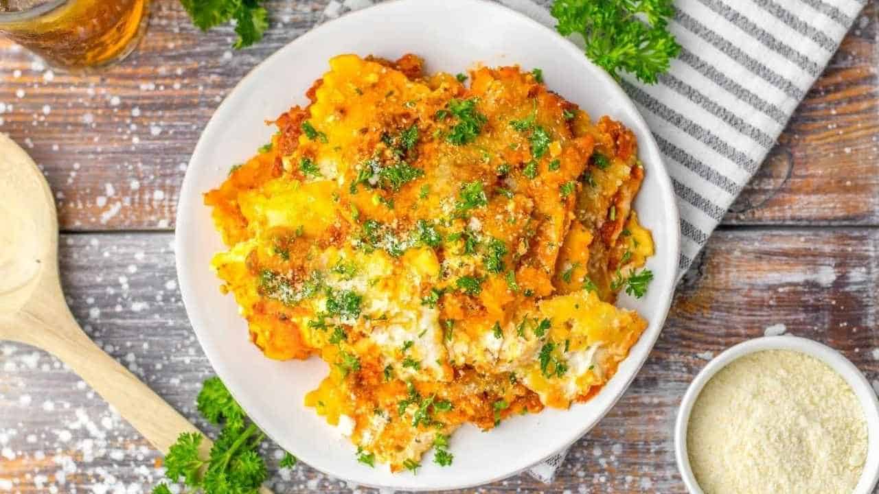 A plate of baked lasagna topped with melted cheese and chopped parsley, placed on a wooden table beside a bowl of grated cheese and a striped napkin.