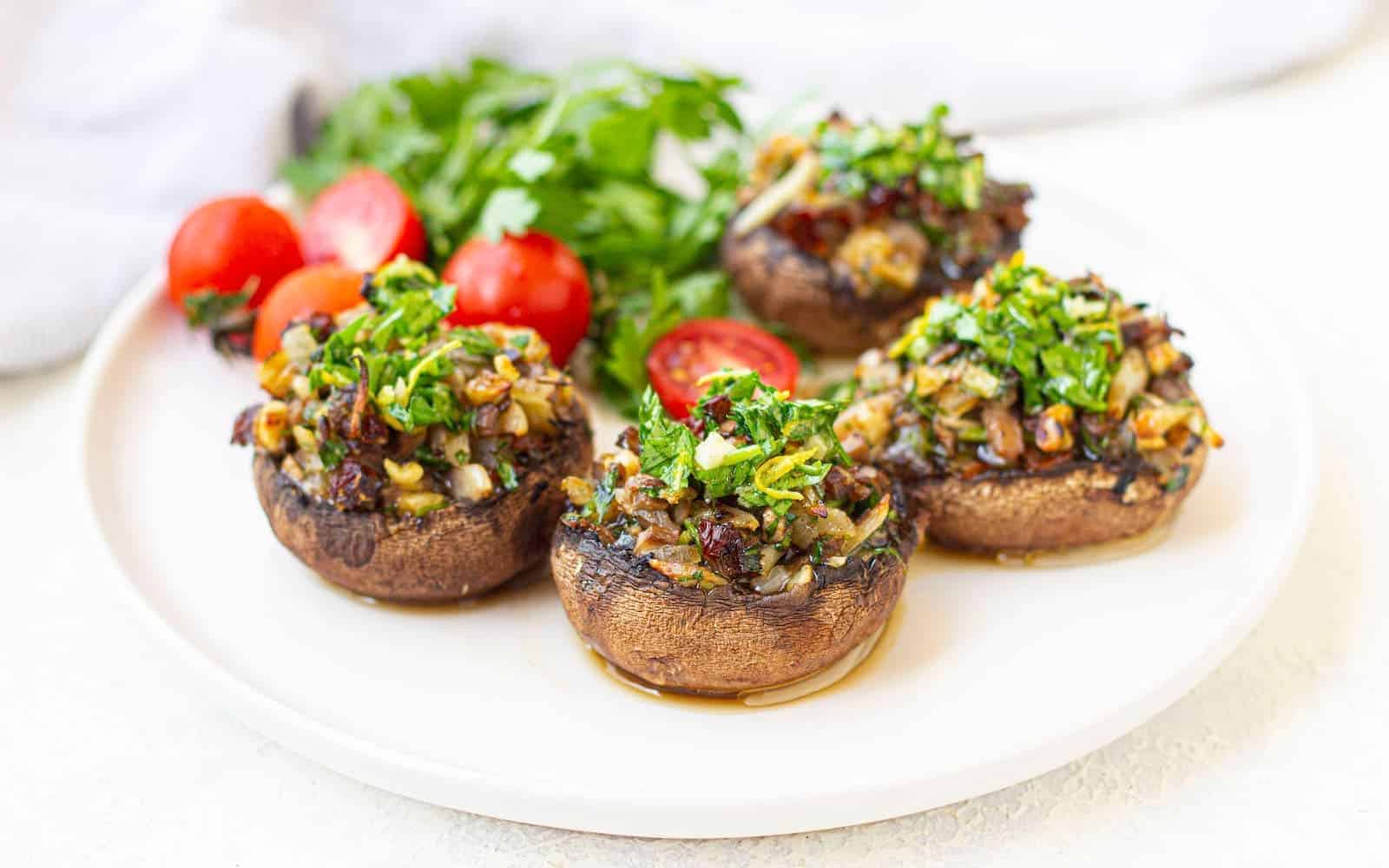 Four stuffed mushrooms topped with chopped herbs are arranged on a white plate, accompanied by cherry tomatoes and fresh parsley.