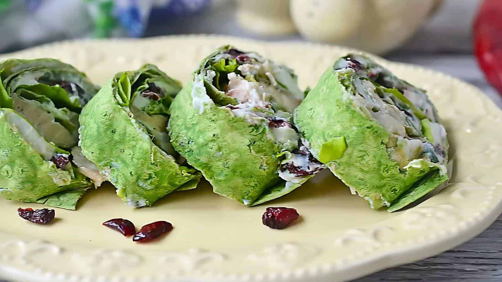Four spinach tortilla wraps with a creamy filling and dried cranberries are arranged on a yellow plate, with a few cranberries scattered nearby.