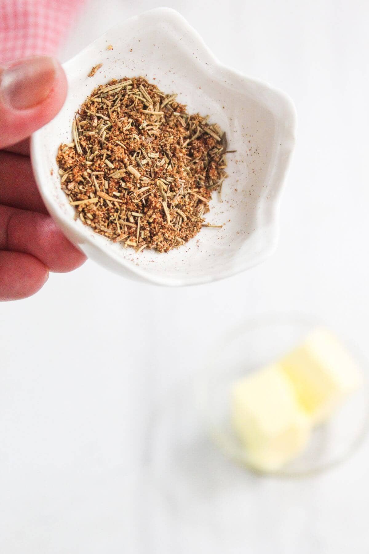 A hand holds a small white dish containing a blend of dried herbs and spices, with a bowl of butter in the blurred background.