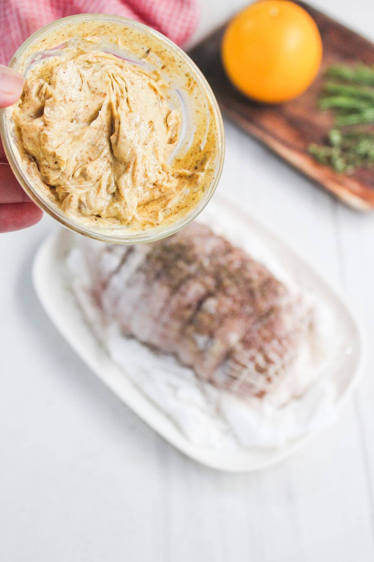 A hand holds a bowl of seasoned butter above a platter with a raw, seasoned meat roast; an orange and herbs are on a cutting board in the background.
