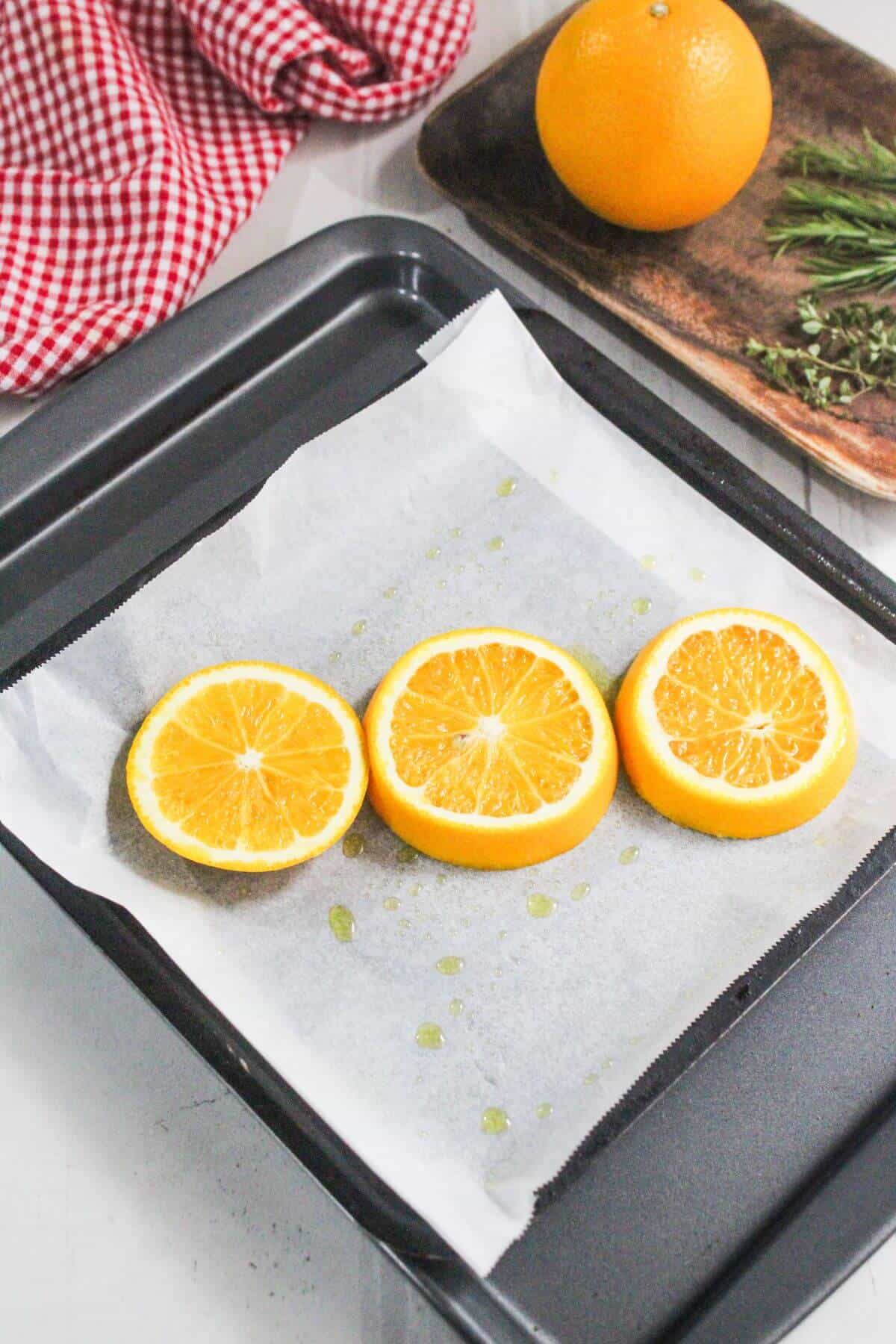 Three orange slices on parchment paper in a baking tray, with oil drizzles visible. A whole orange, rosemary, and a red-checked cloth are in the background.